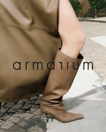 A woman's leg is shown wearing a knee-high, light brown leather boot, and the background includes cobblestones and a hint of greenery.