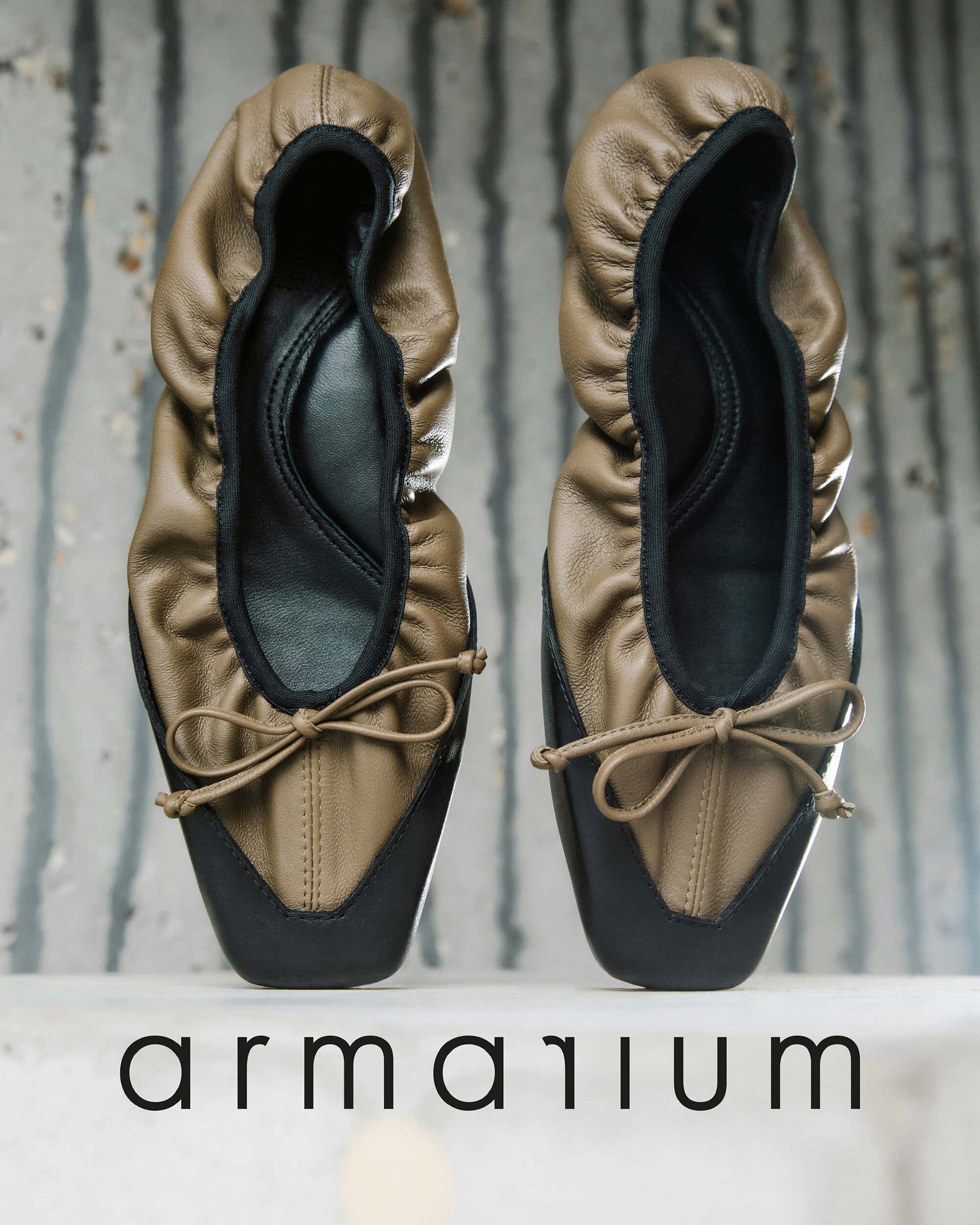 Studio shot of a pair of Armarium women's ballet shoes with a tan and black leather upper and tan bows, set against a textured background.