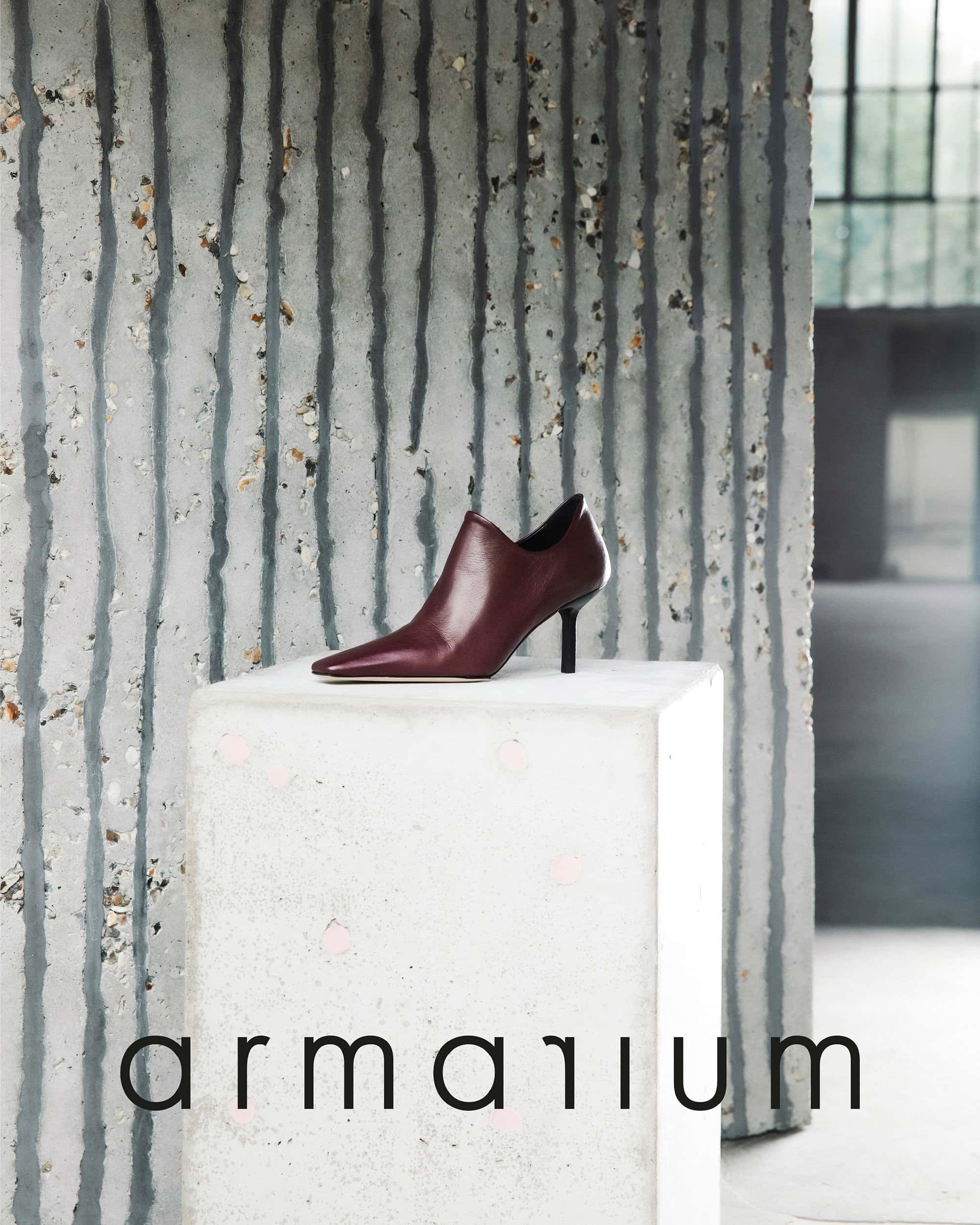 Maroon heeled ankle boot on a white cube against a textured grey wall, featuring the brand 'Armarium' at the base. Fashion-forward studio shot.
