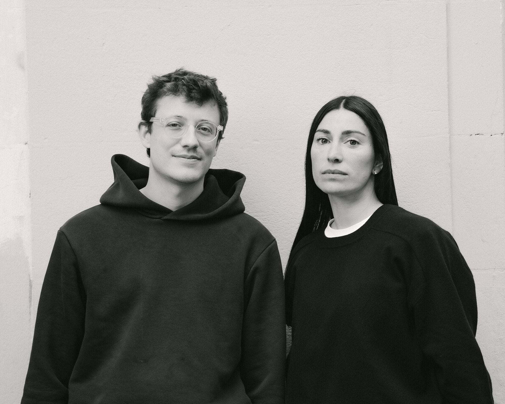 Monochrome portrait of a smiling man in glasses and a hooded sweatshirt standing next to a woman in a long-sleeved shirt against a wall.