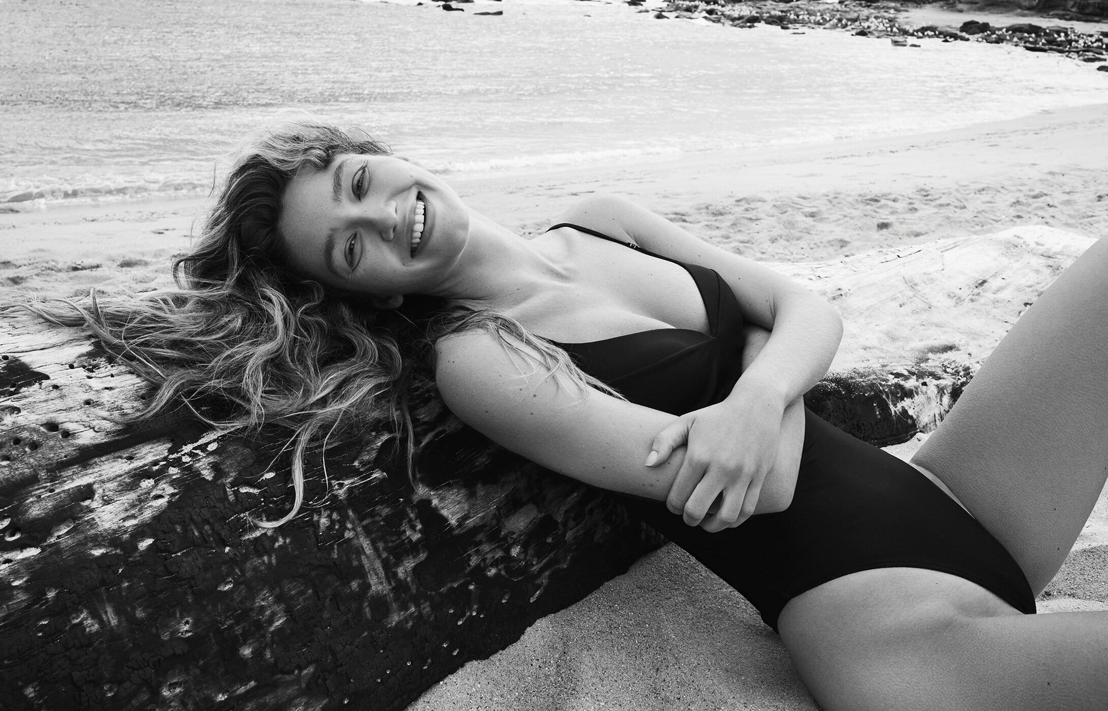 Woman in black swimsuit lying on driftwood at beach, smiling, black and white. Relaxation, swimwear, beach landscape.