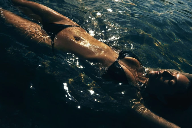 Woman in black bikini swimming in water with sunlight reflecting off of her body and creating sparkling reflections on the surface.