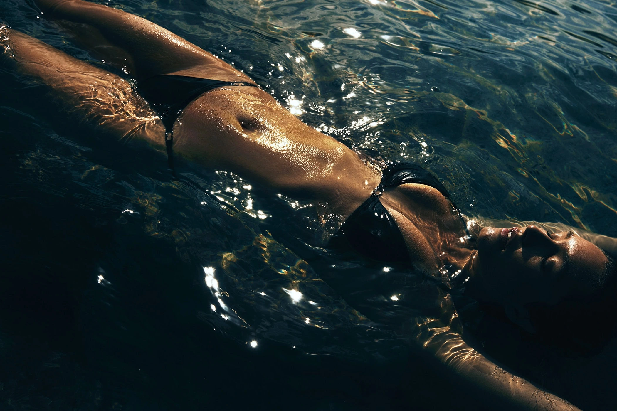 Model floating in dark water wearing a black bikini with shimmering sunlight reflections on wet skin