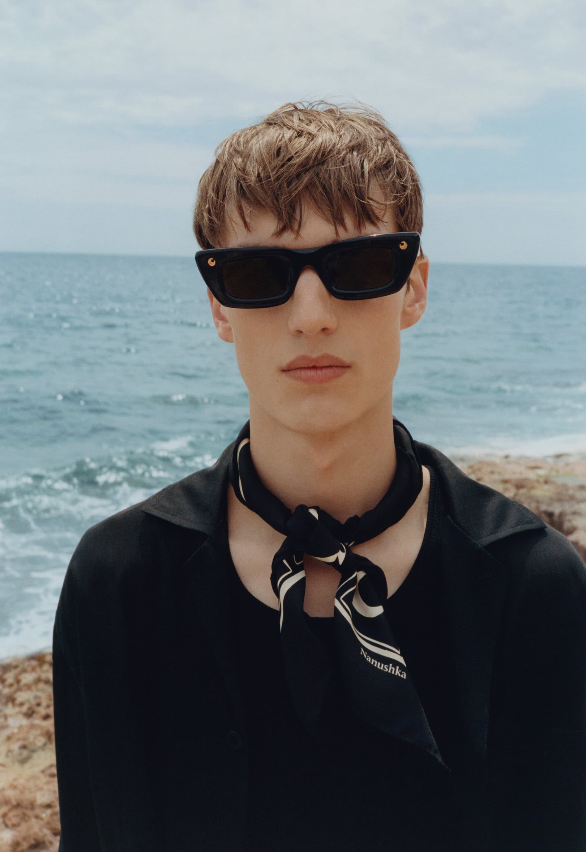 A close-up shot captures a male model wearing sunglasses and a patterned scarf against a blurred seascape under a bright sky.