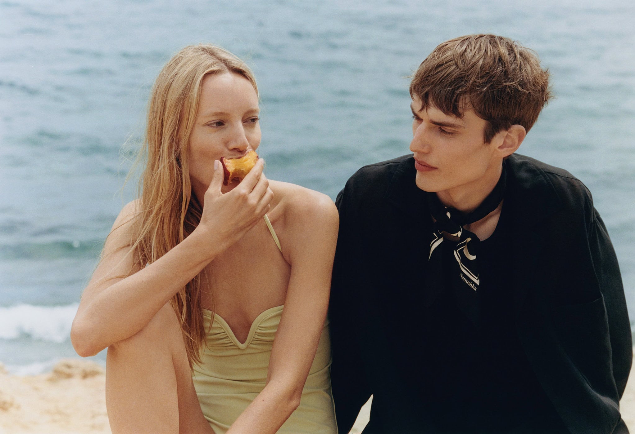 Young couple relaxes on a rocky beach with a light-blue ocean backdrop. The woman eats a peach while the man gazes at her.