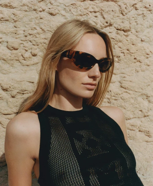 A fair-skinned woman wearing sunglasses and a black patterned top poses against a textured sandstone wall in a close-up studio portrait.