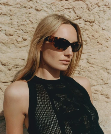A fair-skinned woman wearing sunglasses and a black patterned top poses against a textured sandstone wall in a close-up studio portrait.