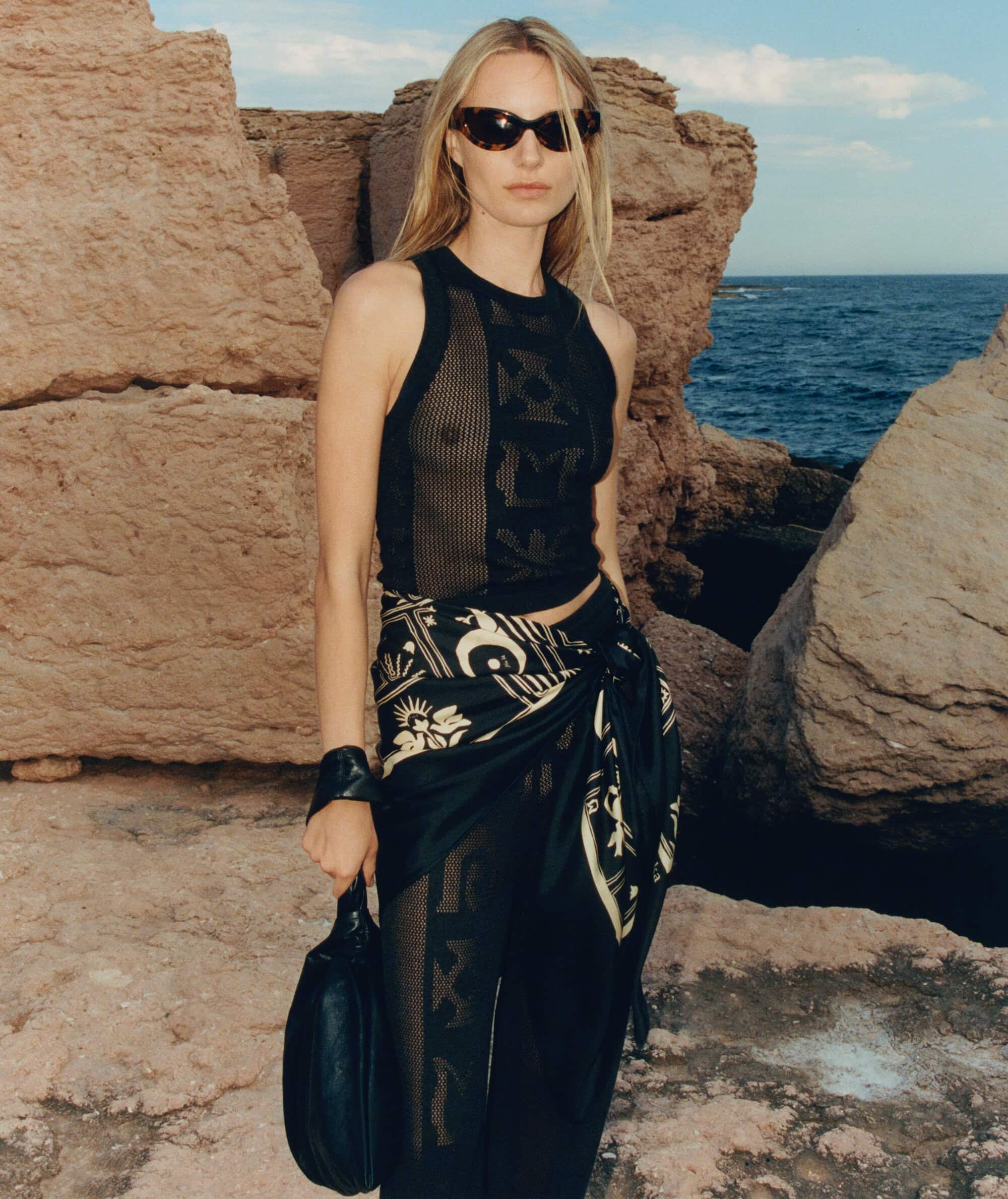 A blonde female fashion model poses outdoors near rocky cliffs wearing a black tank top, patterned pants, sunglasses, and carrying a handbag.