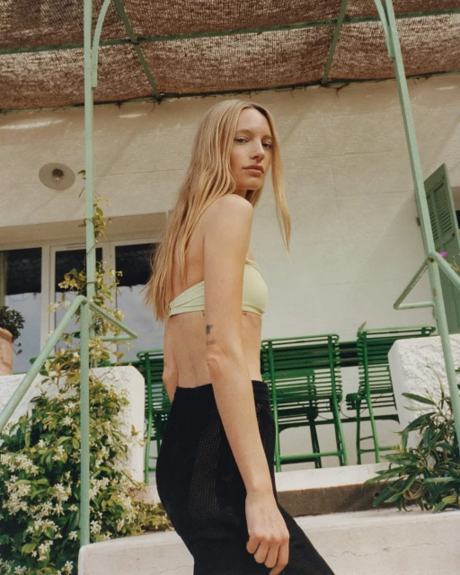 A young adult female poses on a stairwell wearing a pale green bandeau top and black pants, in a sun-drenched outdoor setting.