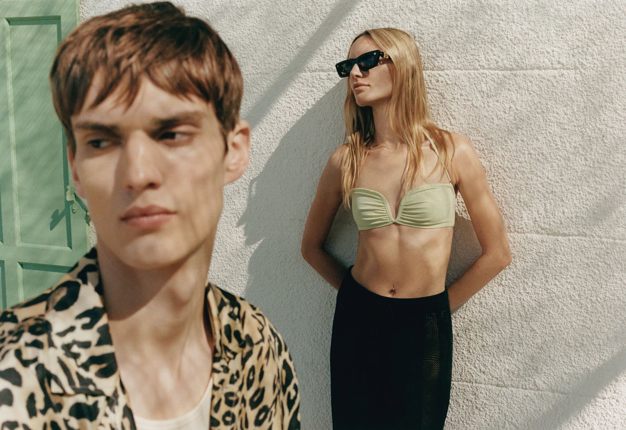 Fashion shot of an adult couple in swimwear and a leopard-print shirt posing outdoors on a sunny day in front of a white wall.