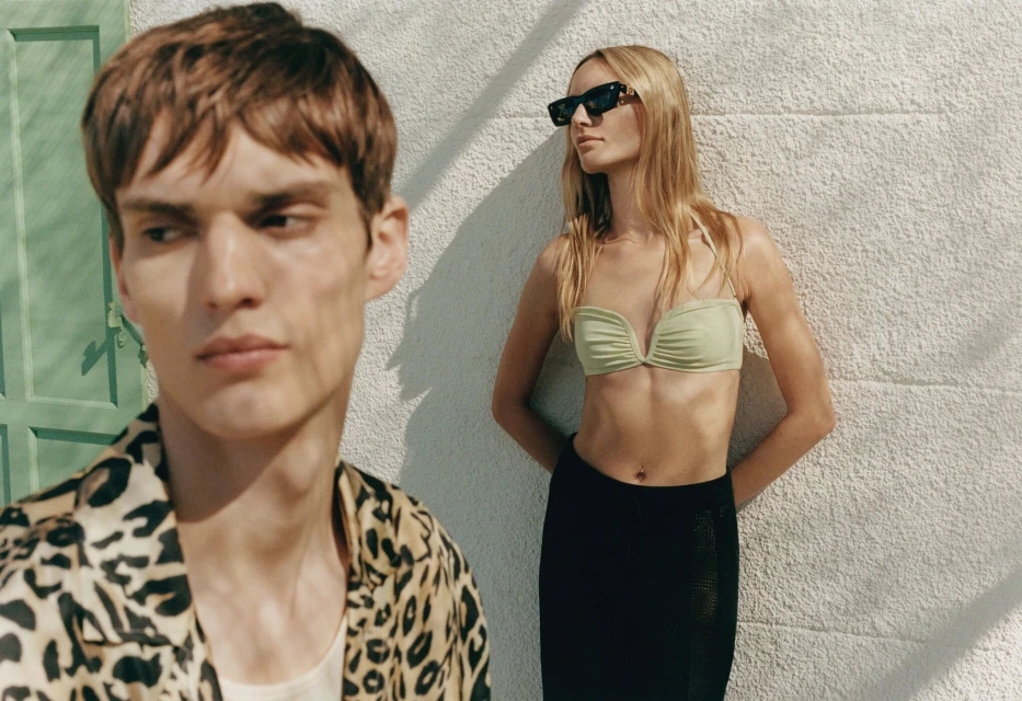 Fashion shot of an adult couple in swimwear and a leopard-print shirt posing outdoors on a sunny day in front of a white wall.