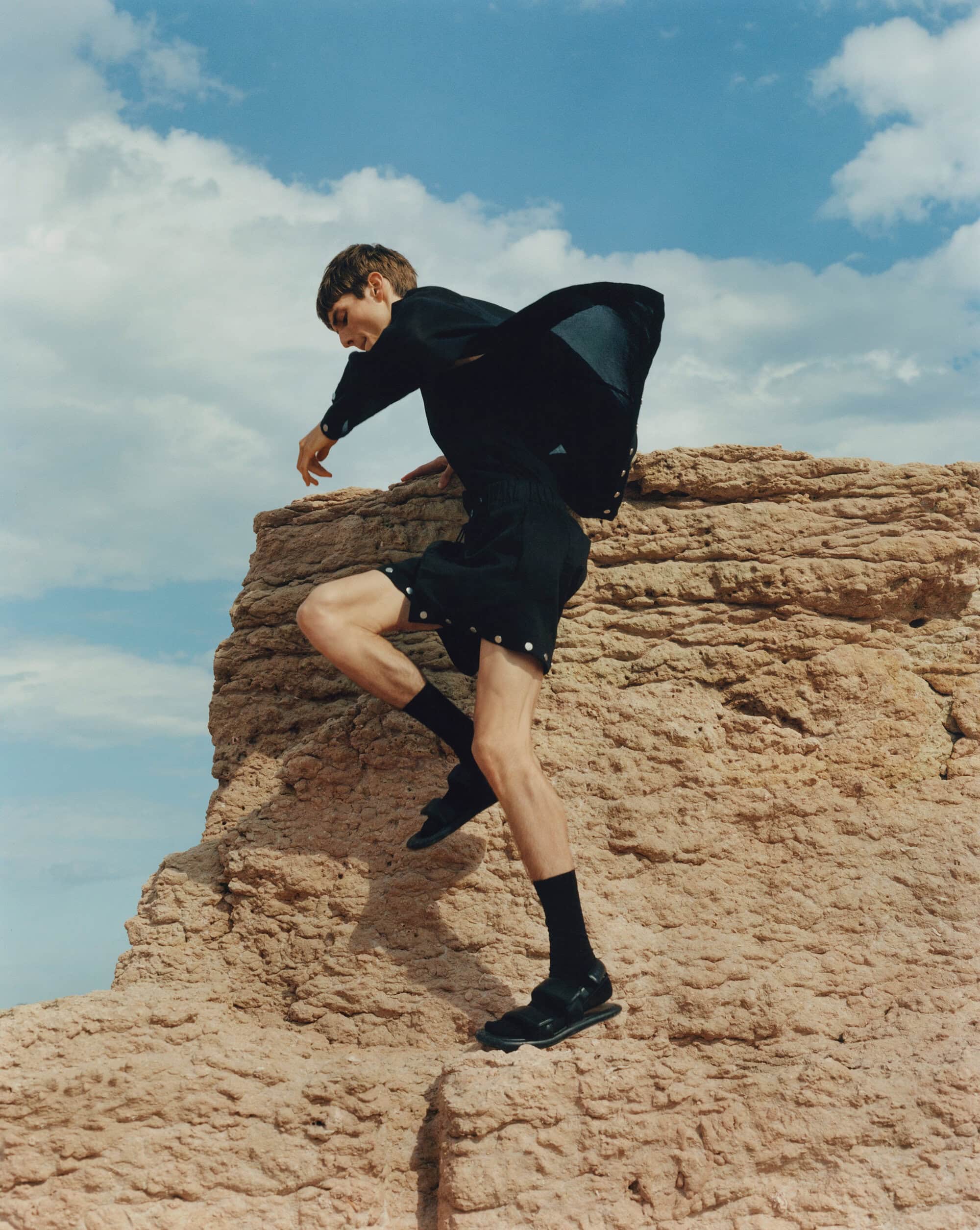 A slender man in dark shorts and a jacket strikes a dynamic pose atop a textured rock formation against a partly cloudy sky.