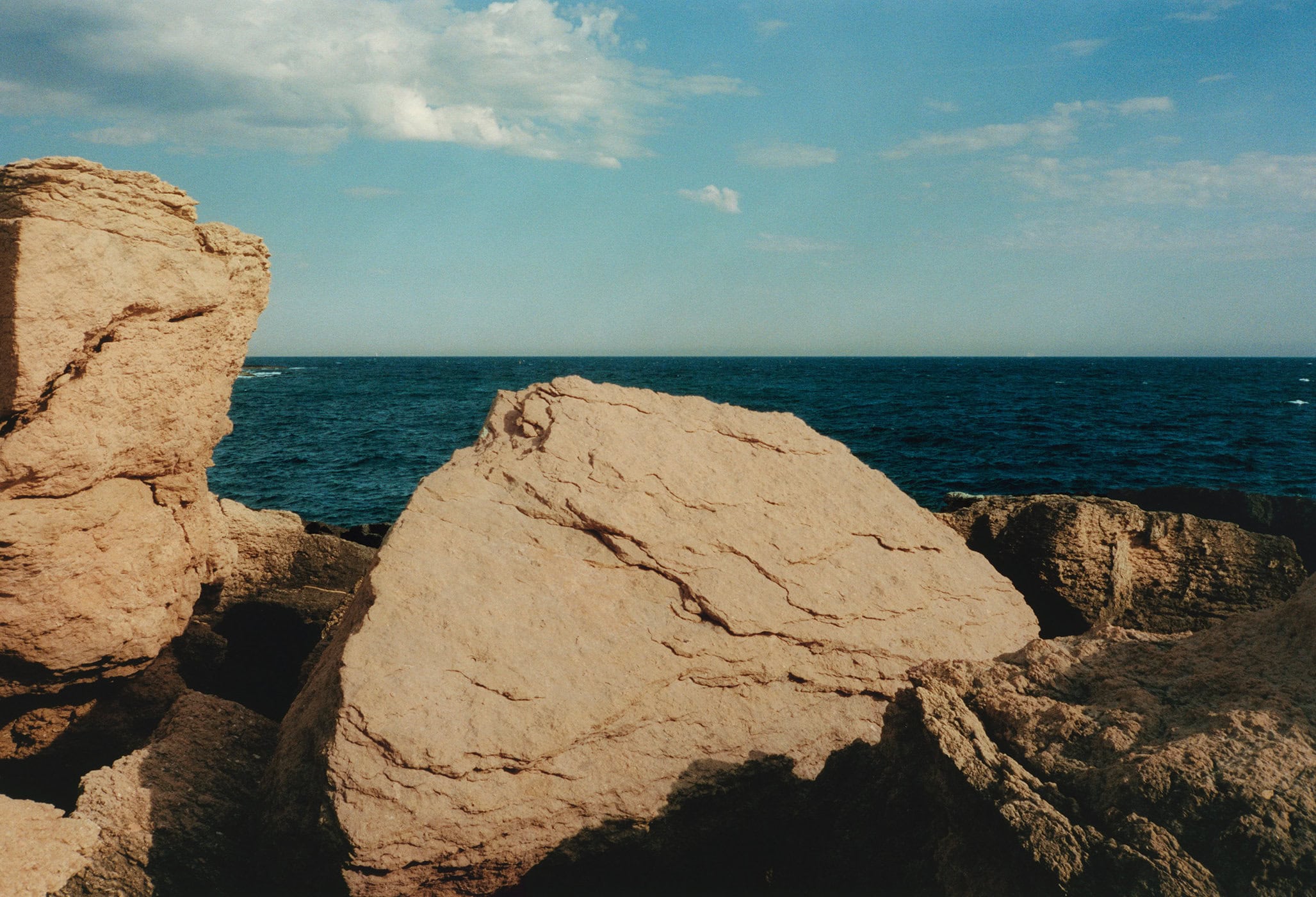 Coastal rock formations meet the ocean under a blue sky with scattered clouds, creating a serene seascape with natural textures.