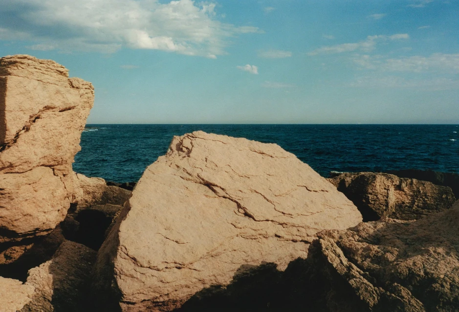 Coastal rock formations meet the ocean under a blue sky with scattered clouds, creating a serene seascape with natural textures.