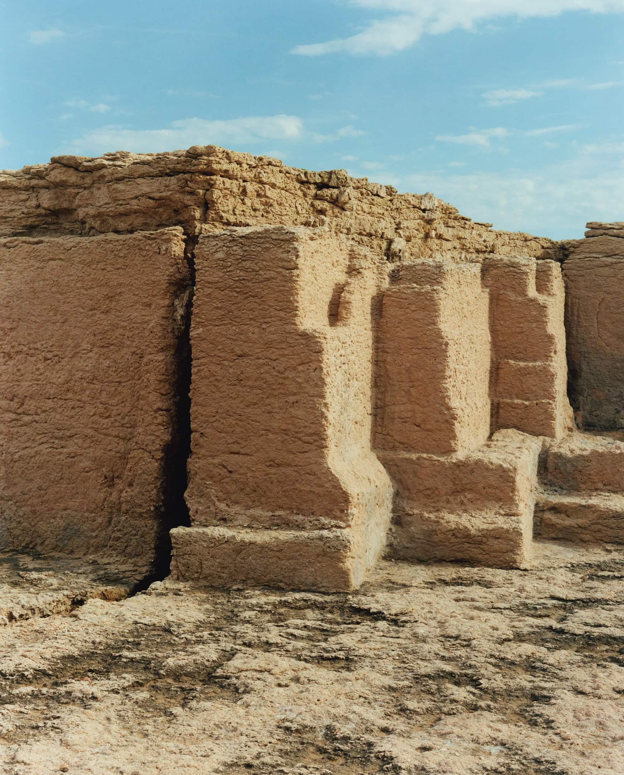 Ruins of a brown stone structure with a wall and columns under a light blue sky with some clouds, landscape orientation.