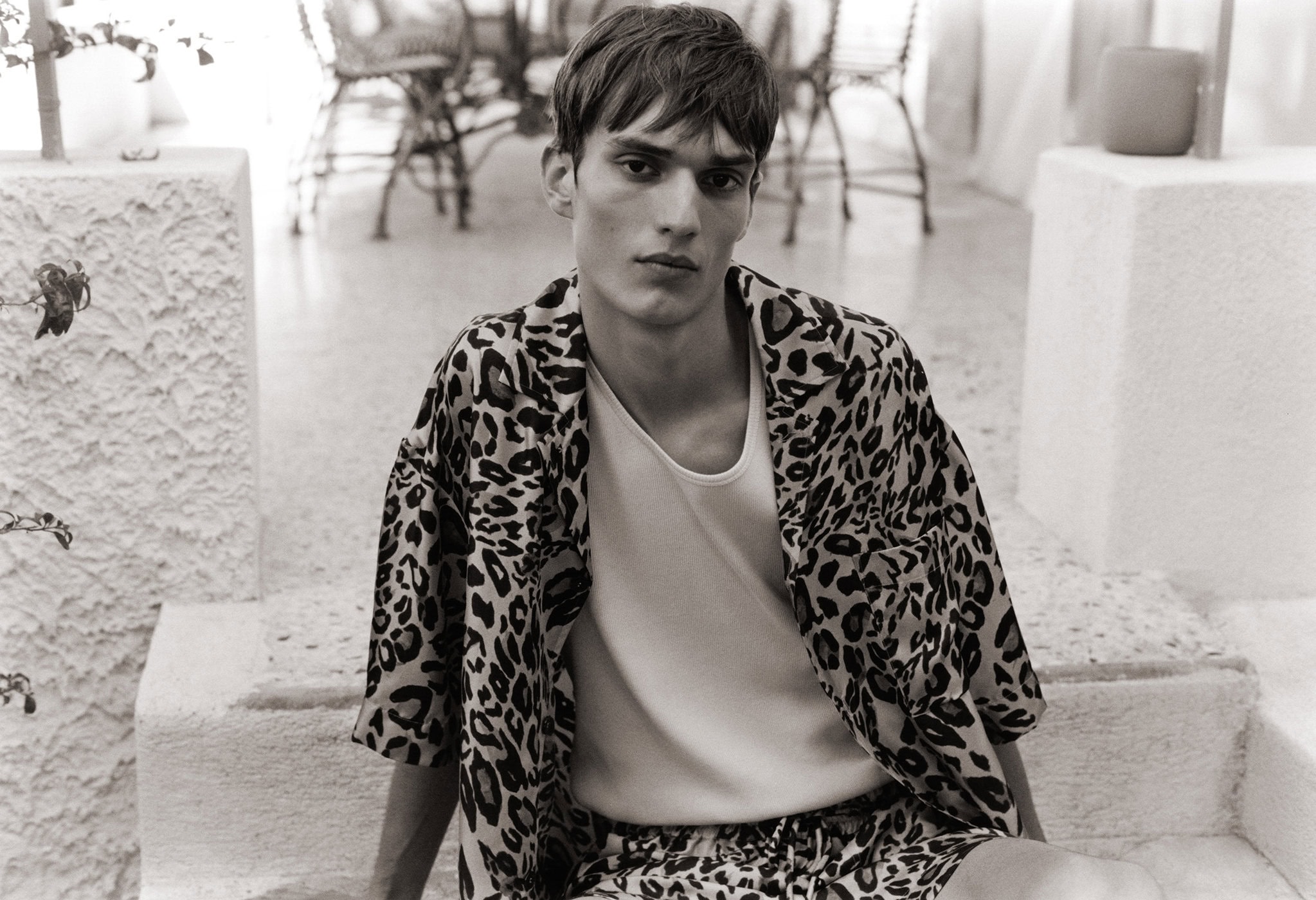 Monochrome portrait of a young man in animal print shirt and shorts, posing on white concrete steps in an indoor/outdoor setting.