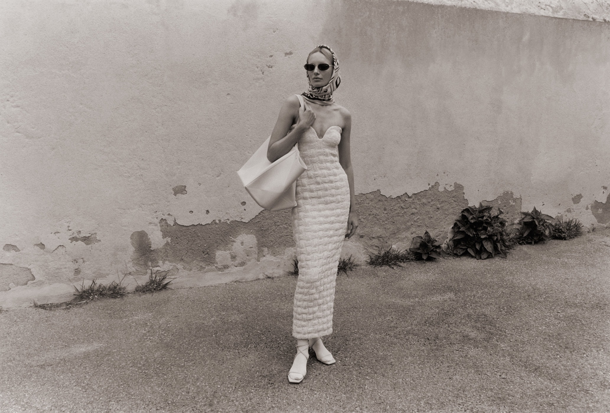 Monochrome shot of a woman in a textured dress and scarf, carrying a tote bag, standing in front of a distressed wall.