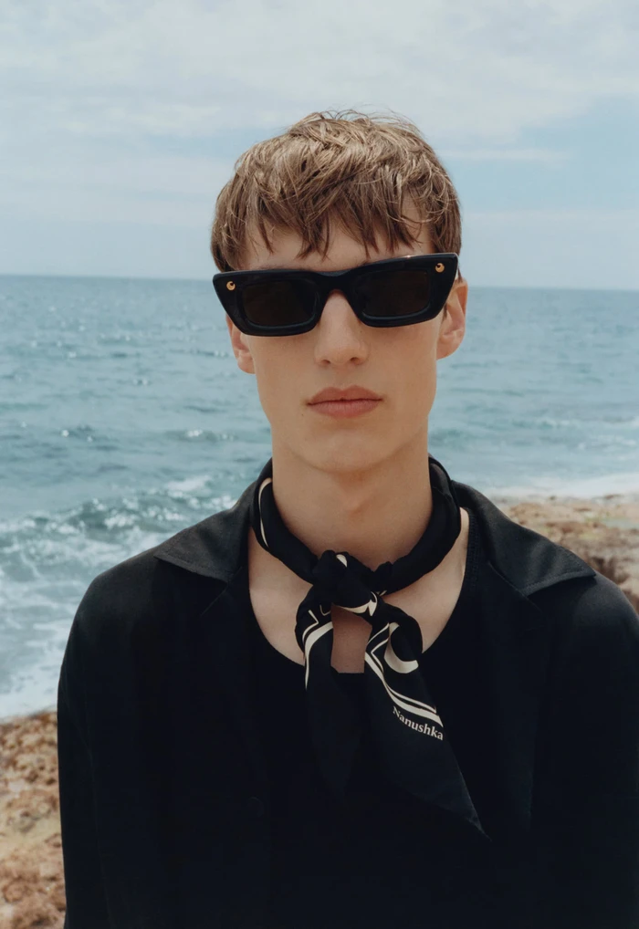 Model wearing black sunglasses and a black shirt with a patterned neck scarf against an ocean backdrop