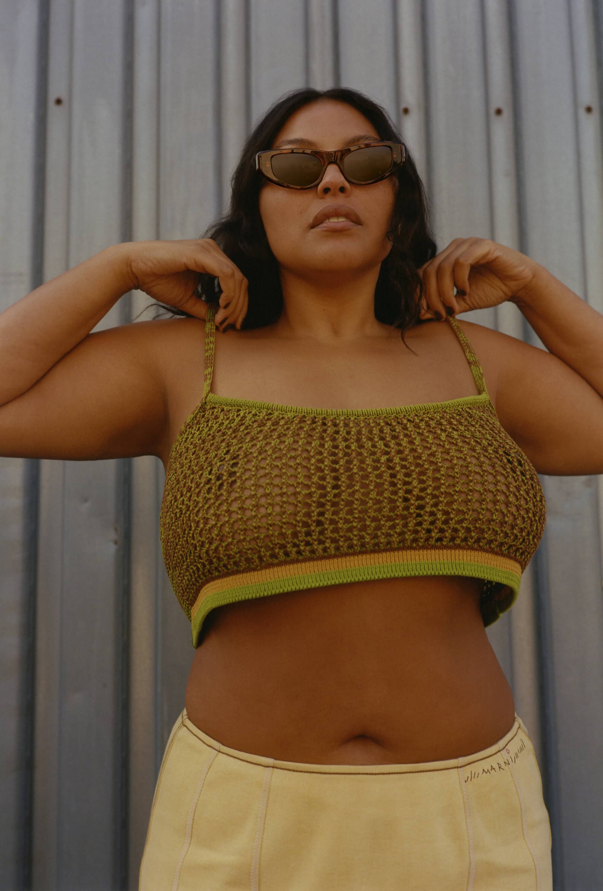 Woman in knit crop top and skirt poses in front of corrugated metal wall wearing sunglasses, looking directly at the camera.