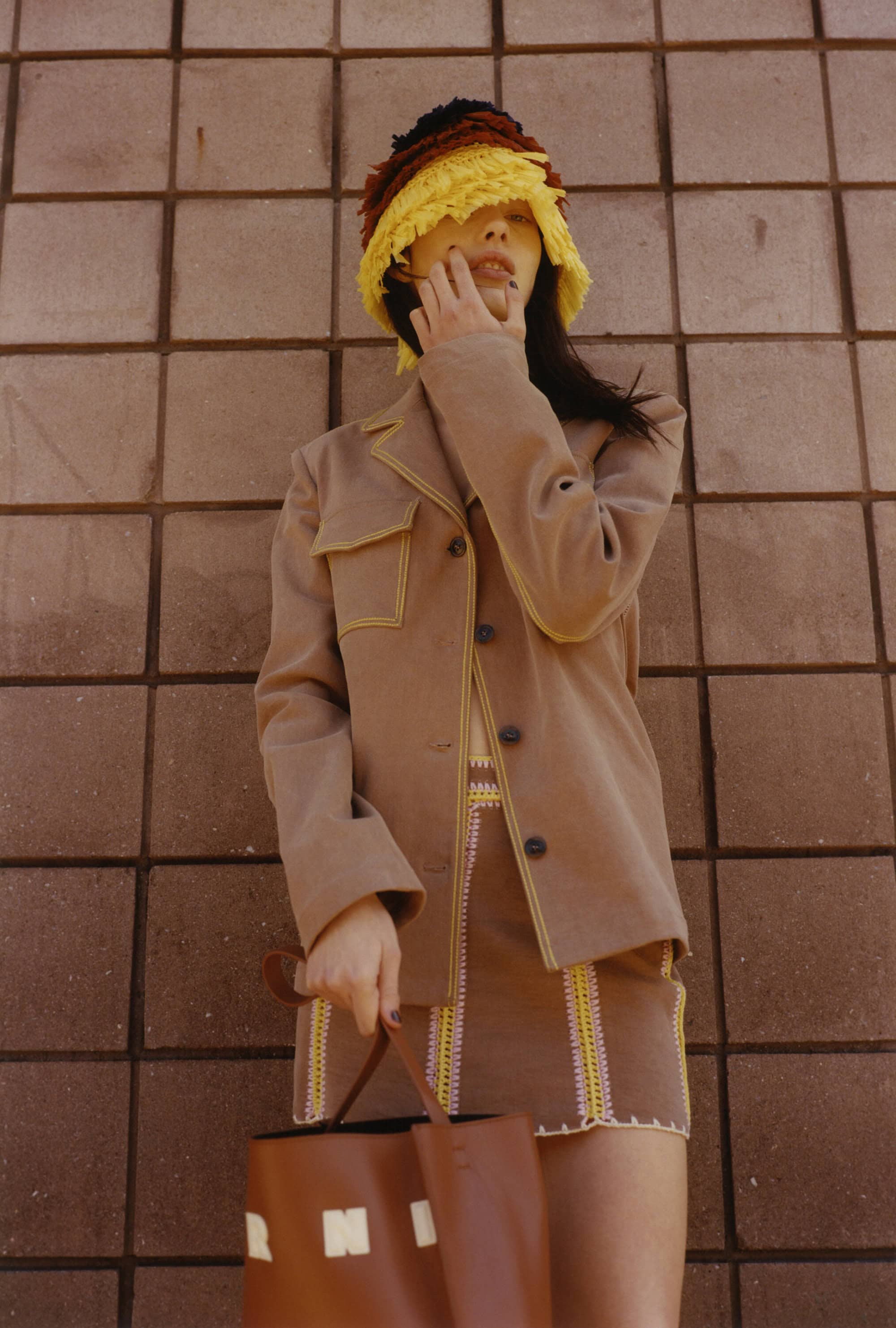 Woman in tan jacket and mini skirt poses against tile wall, holding brown bucket bag and wearing colorful crochet hat in a fashion shot.