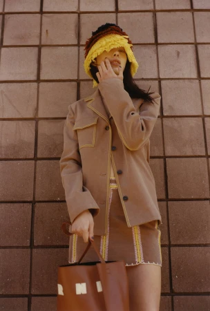 Woman in tan jacket and mini skirt poses against tile wall, holding brown bucket bag and wearing colorful crochet hat in a fashion shot.
