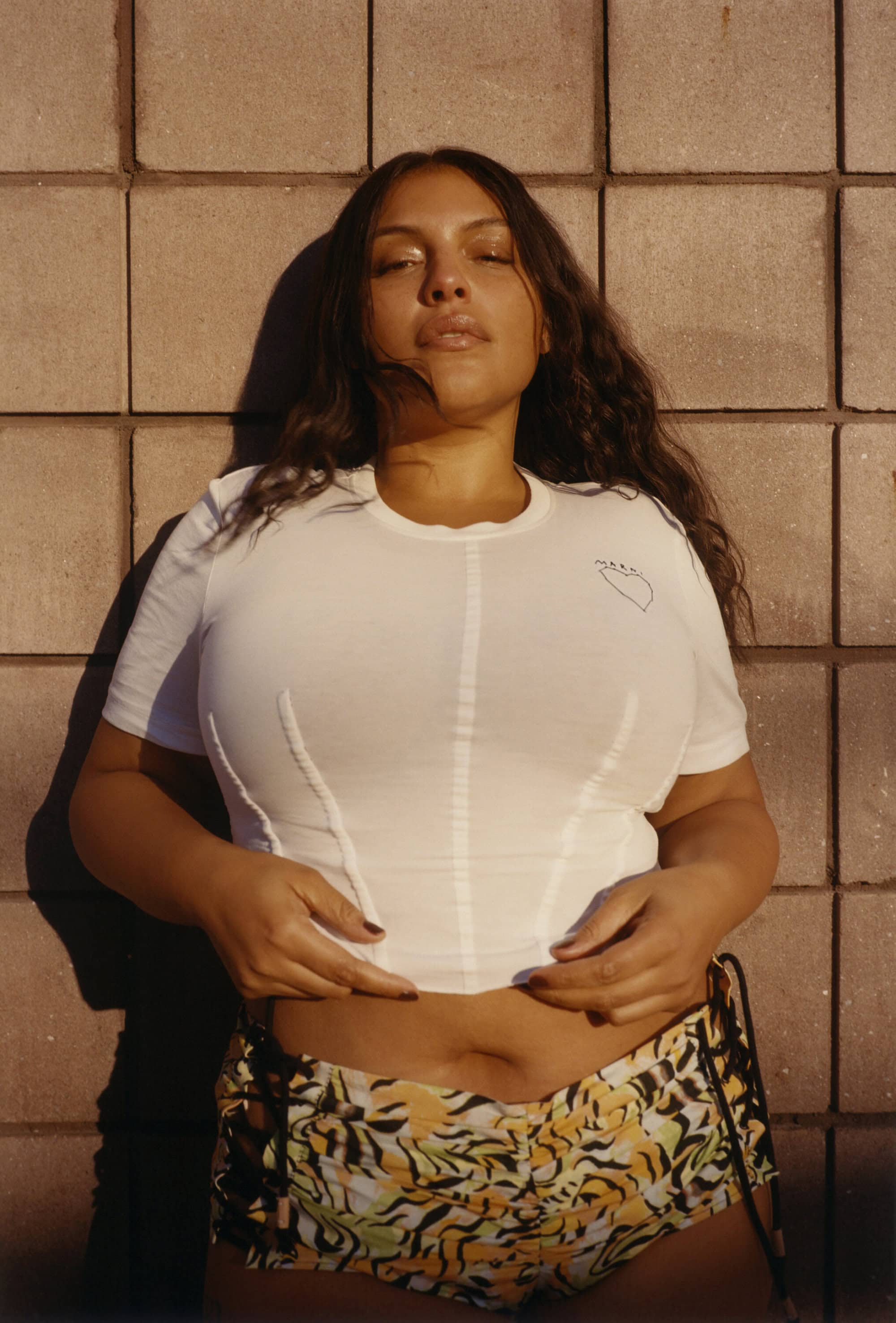 A woman with long brown hair stands in front of a wall of brick. She is wearing a white shirt and patterned shorts.