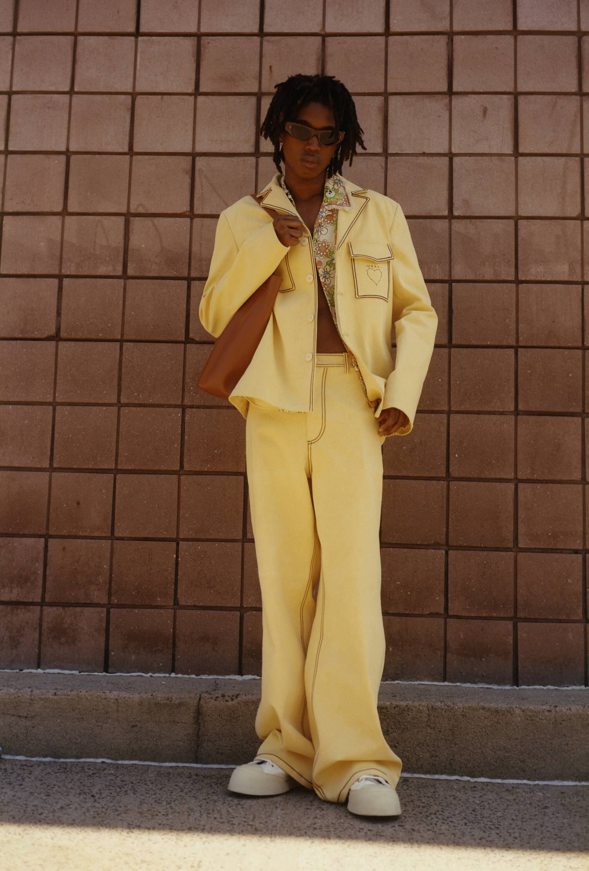 An adult person with dreadlocks in a yellow suit and sunglasses carries a brown bag, posing against a square brick wall outdoors.