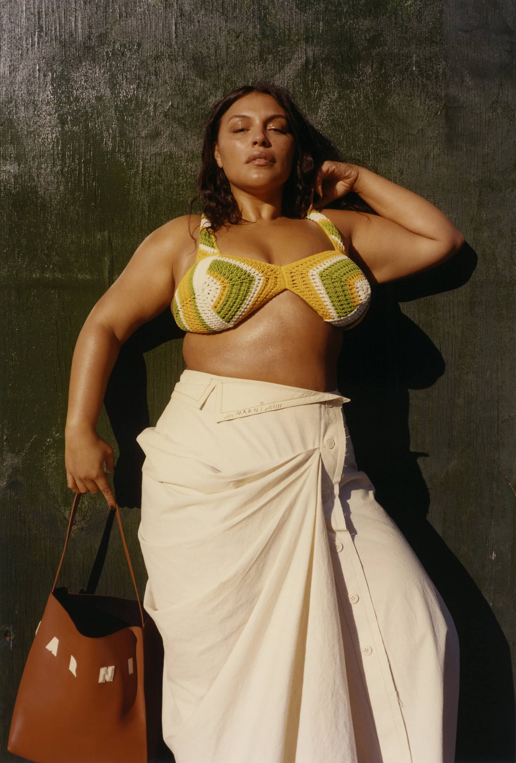 Woman wearing a crochet bikini top and a skirt holding a bag poses against a textured background in an eye-level portrait shot.