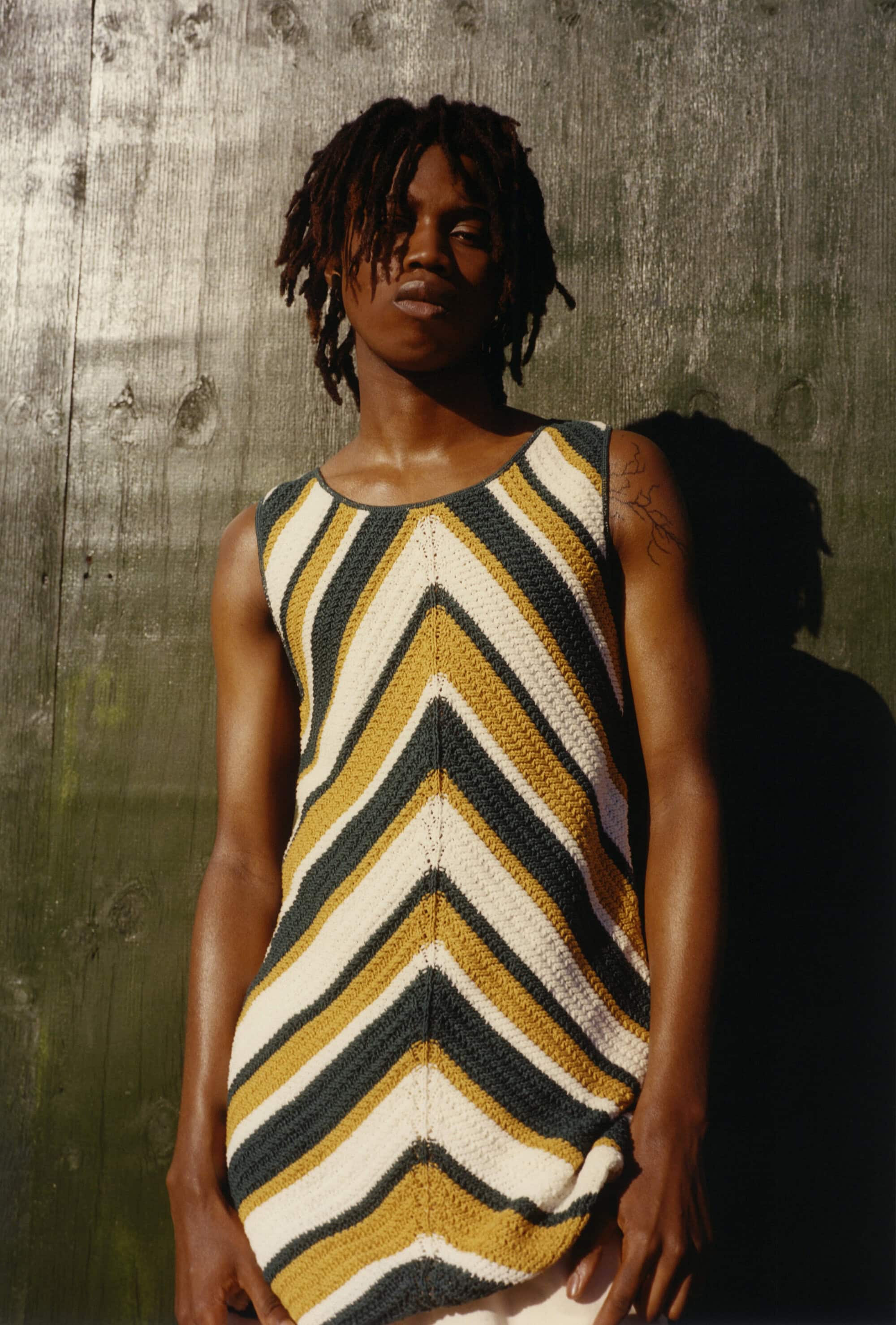 Young adult poses in a sleeveless, striped knit dress, standing outdoors against a weathered wooden wall. Natural lighting, portrait style.