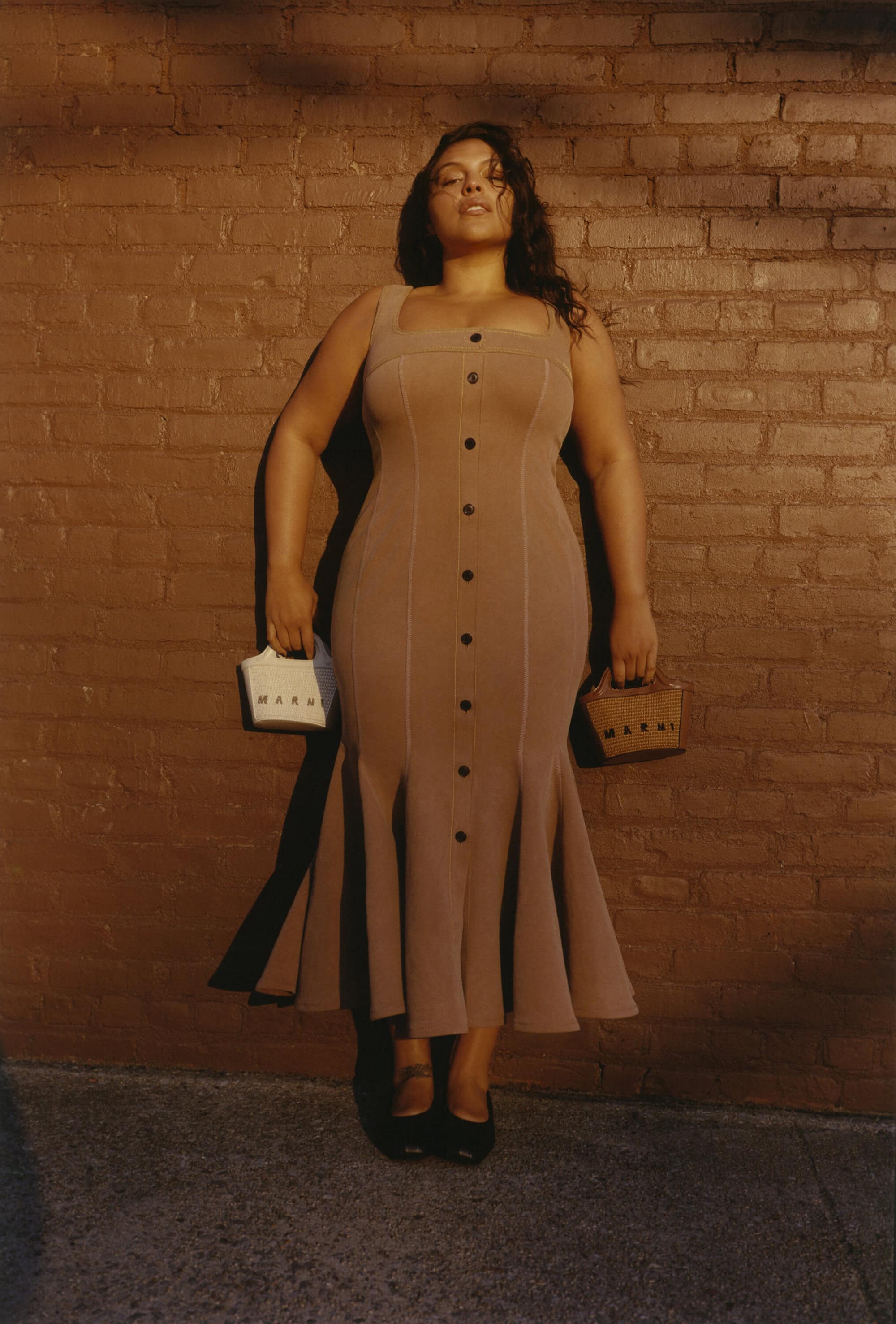 Adult woman poses in front of a brick wall, wearing a fitted dress and black flats, while holding two small bags.