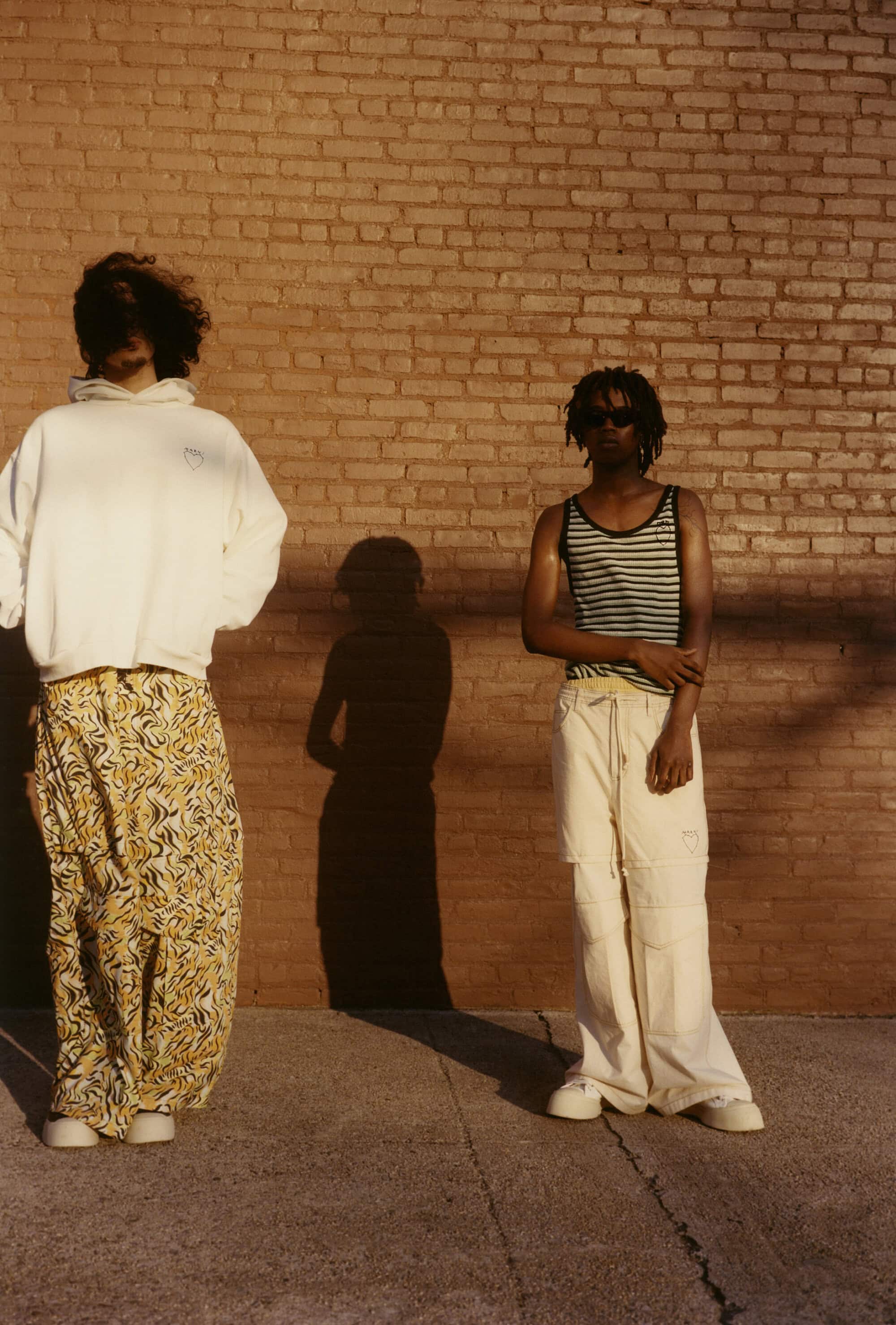 Two people pose in loose-fitting clothing against a brick wall in warm sunlight, casting long shadows on the ground. Fashion-forward style.