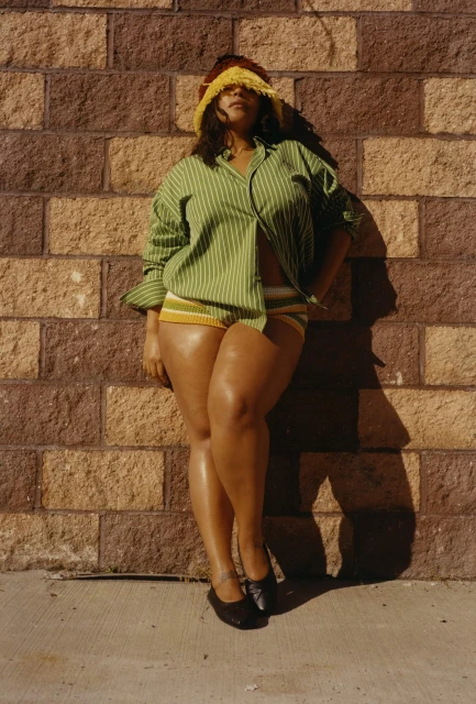 A woman in a green striped shirt and yellow shorts poses against a brick wall, wearing a yellow fringe hat obscuring her eyes.