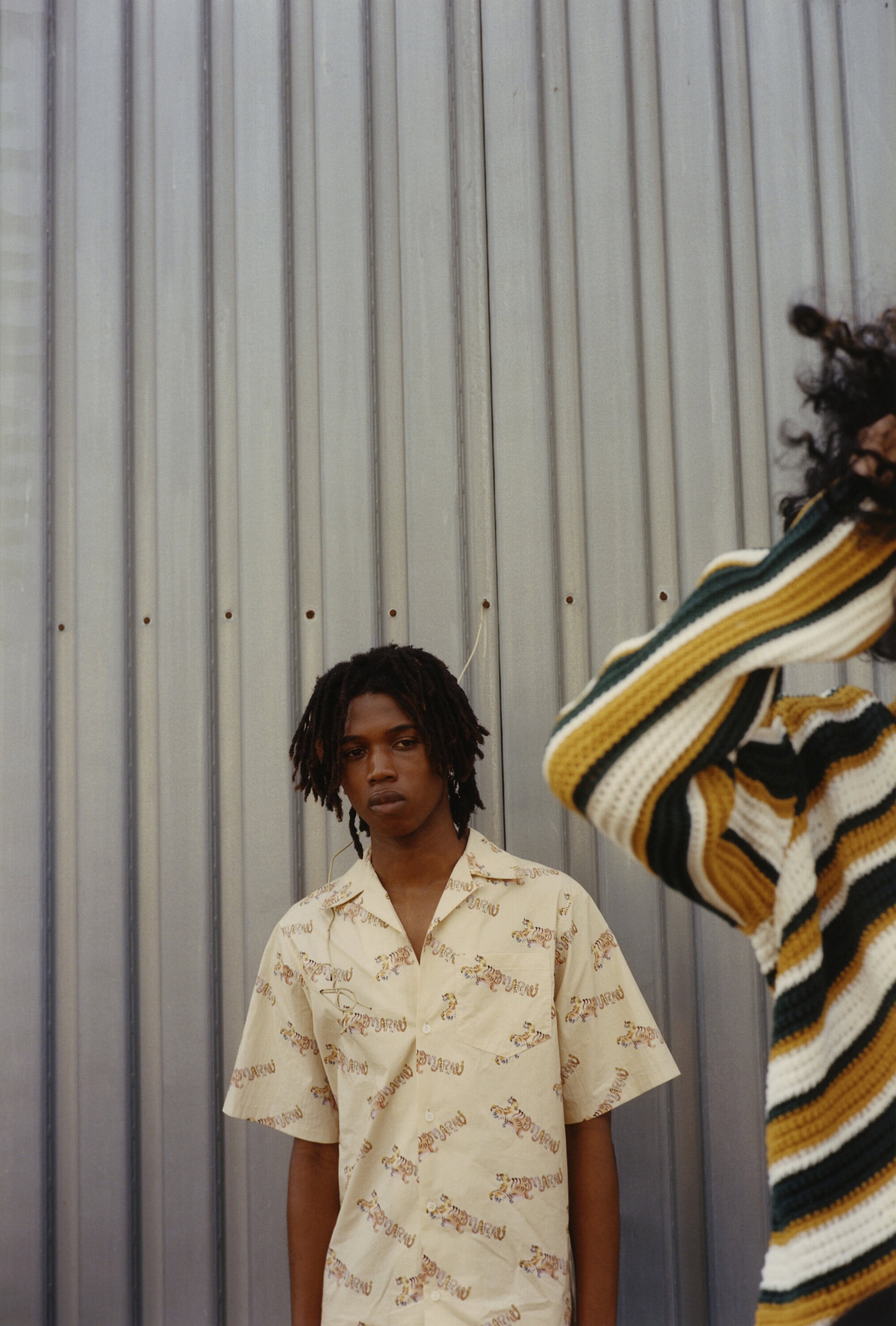 Two African-descent young adults pose in front of a corrugated metal wall. One wears a patterned shirt, the other a striped sweater.