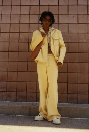 Model wearing a yellow suit jacket and trousers holding a brown shoulder bag against a tiled wall