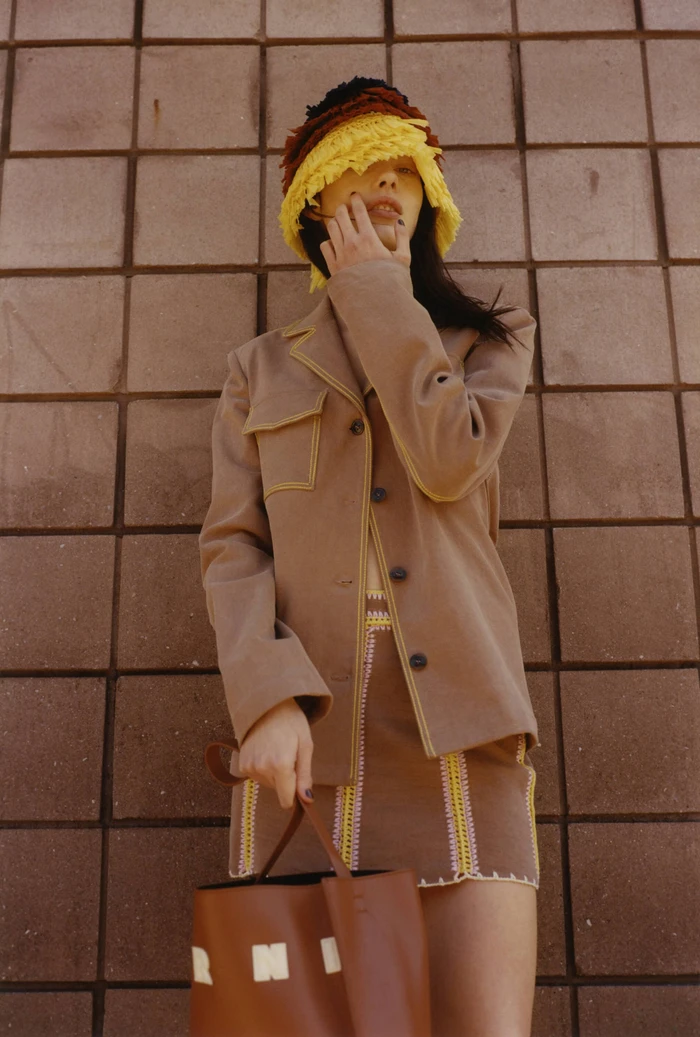 Model Paloma Elsesser in a brown jacket and fringed hat holding a Marni bag against a tiled wall