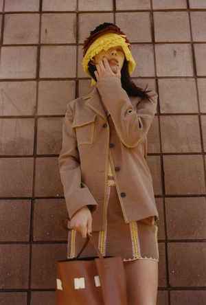 Model Paloma Elsesser in a brown jacket and fringed hat holding a Marni bag against a tiled wall