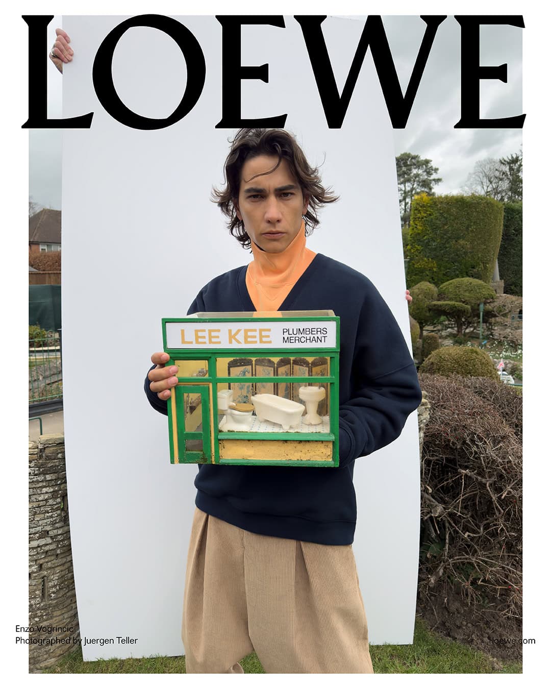 Fashion editorial featuring a young man holding a miniature plumber shop, standing outdoors with a Loewe logo backdrop and scenic elements.