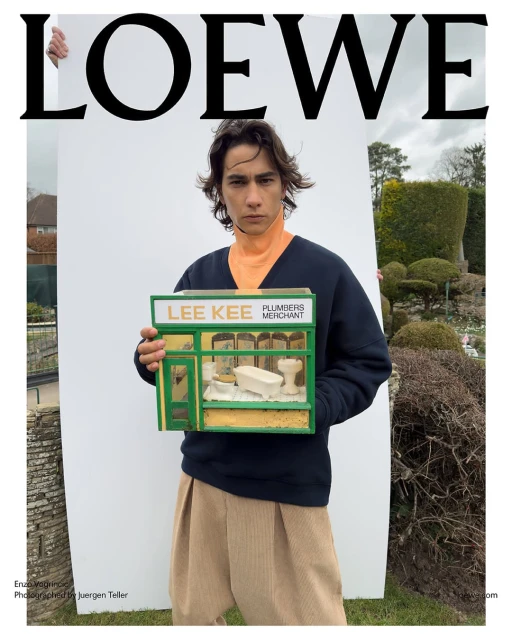 Fashion editorial featuring a young man holding a miniature plumber shop, standing outdoors with a Loewe logo backdrop and scenic elements.