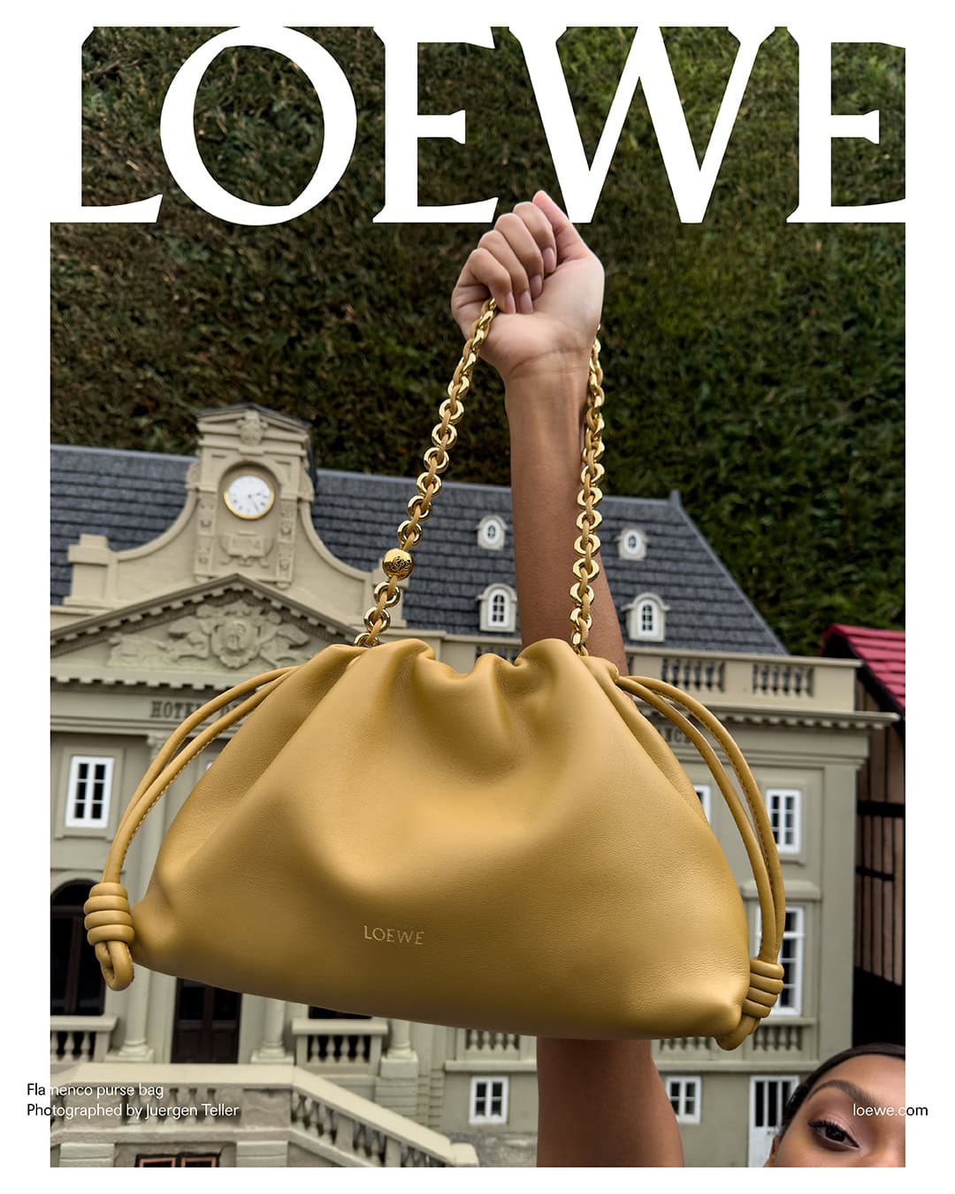 Tan-colored Loewe drawstring purse with gold chain handle held up against a miniature building and green foliage backdrop.