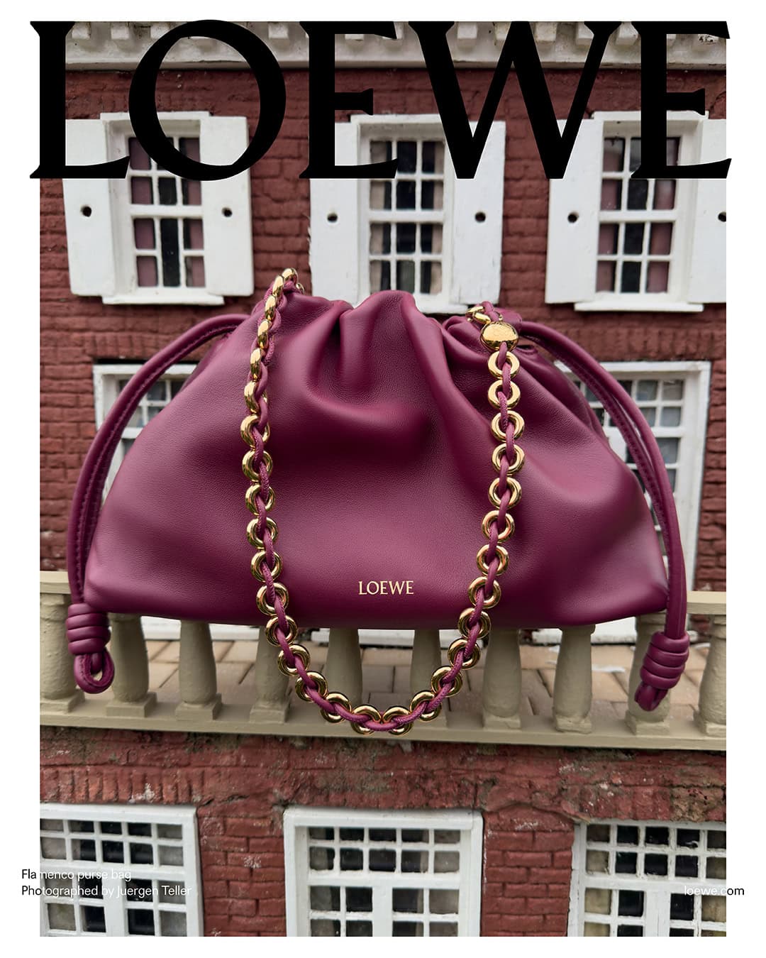 Burgundy Loewe pouch bag with gold chain on miniature balcony against a brick building with white shutters in an indoor setting.