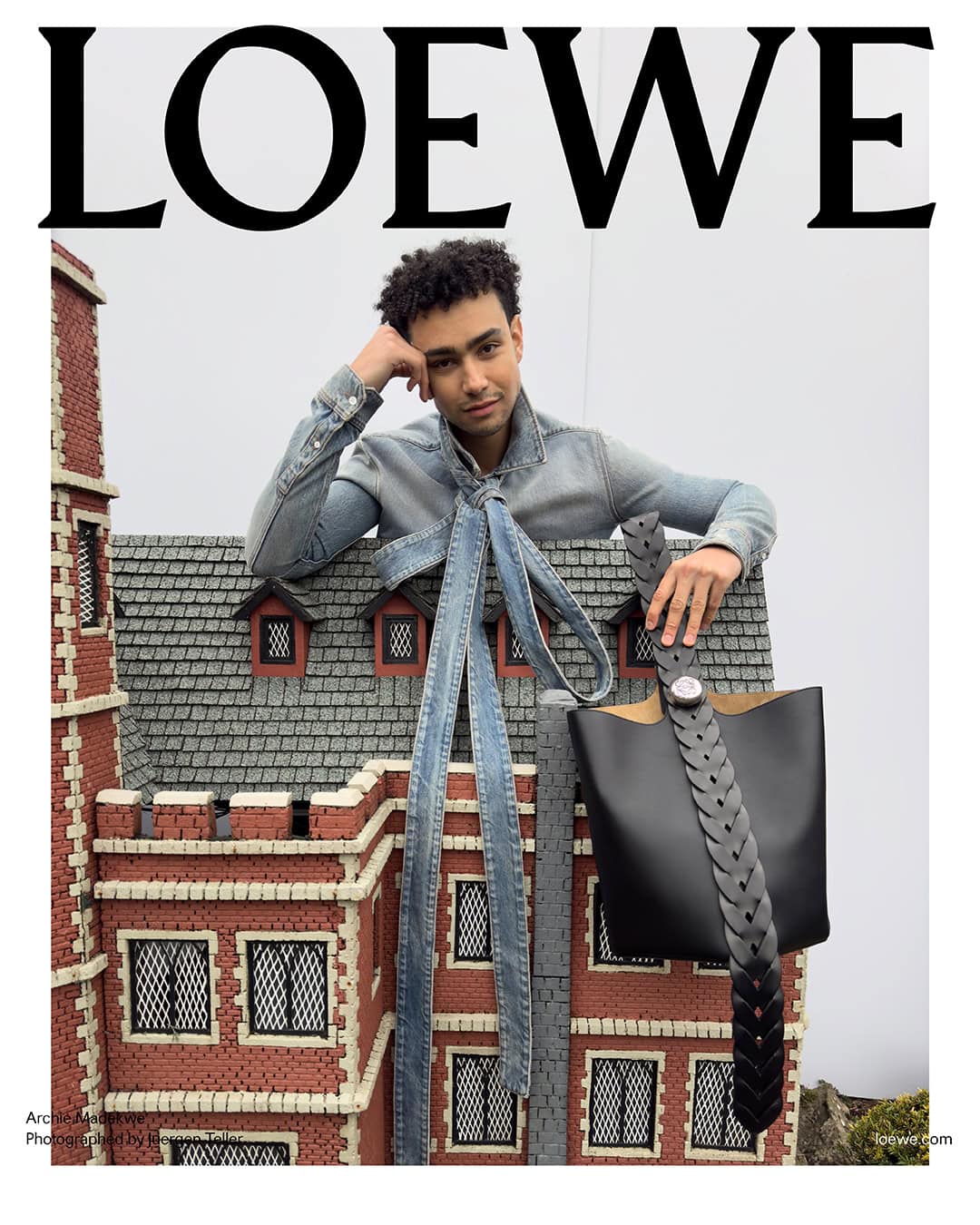 Fashion editorial featuring a person with curly hair in a denim outfit, leaning on a brick building miniature set with a black leather bag.