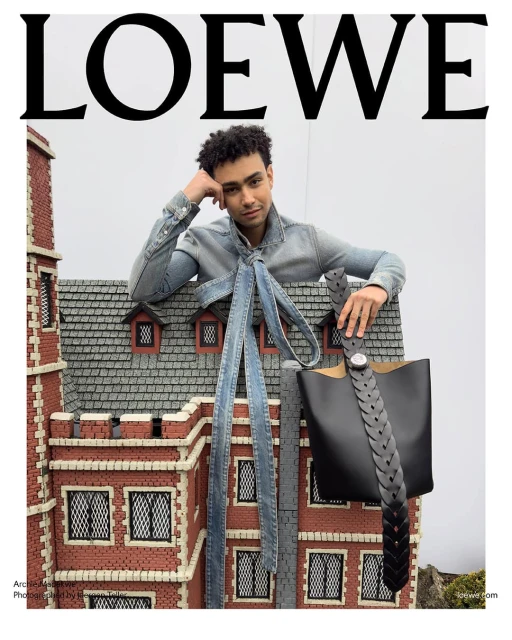 Fashion editorial featuring a person with curly hair in a denim outfit, leaning on a brick building miniature set with a black leather bag.