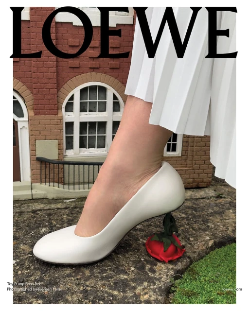 White pump with a rose heel next to a miniature brick building and pleated white skirt. Loewe brand logo.