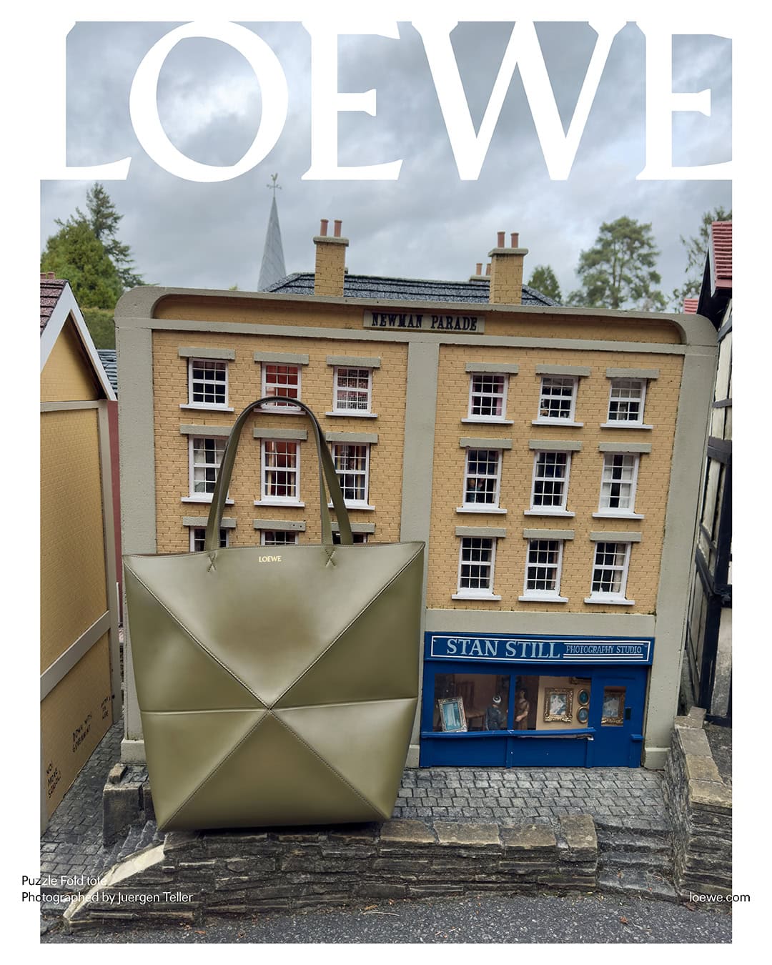 Loewe Puzzle Fold Tote Bag in khaki green stands in front of miniature brick buildings, cobblestone walkway, and overcast sky.