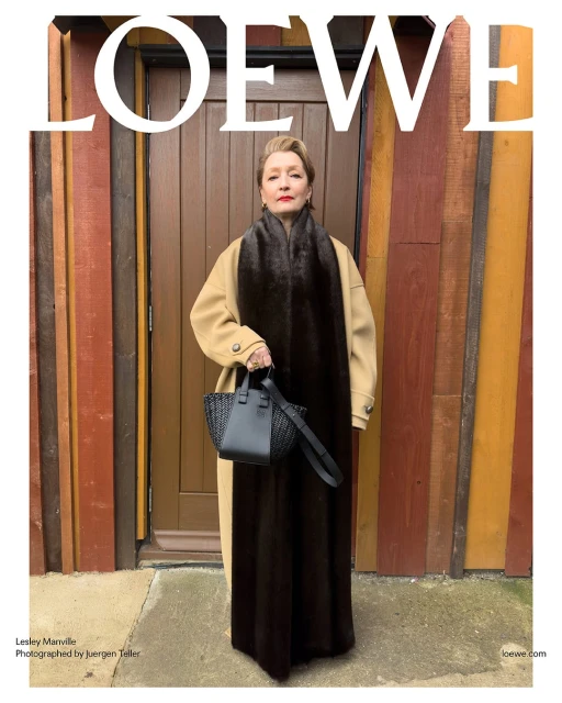 Lesley Manville poses in front of a wooden door wearing a tan coat and brown scarf, holding a black woven handbag with the brand name above.