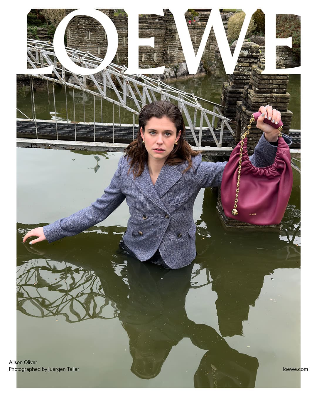 Woman in tweed blazer standing in water, holding a maroon leather purse with gold chain strap, LOEWE logo and the bridge behind her.