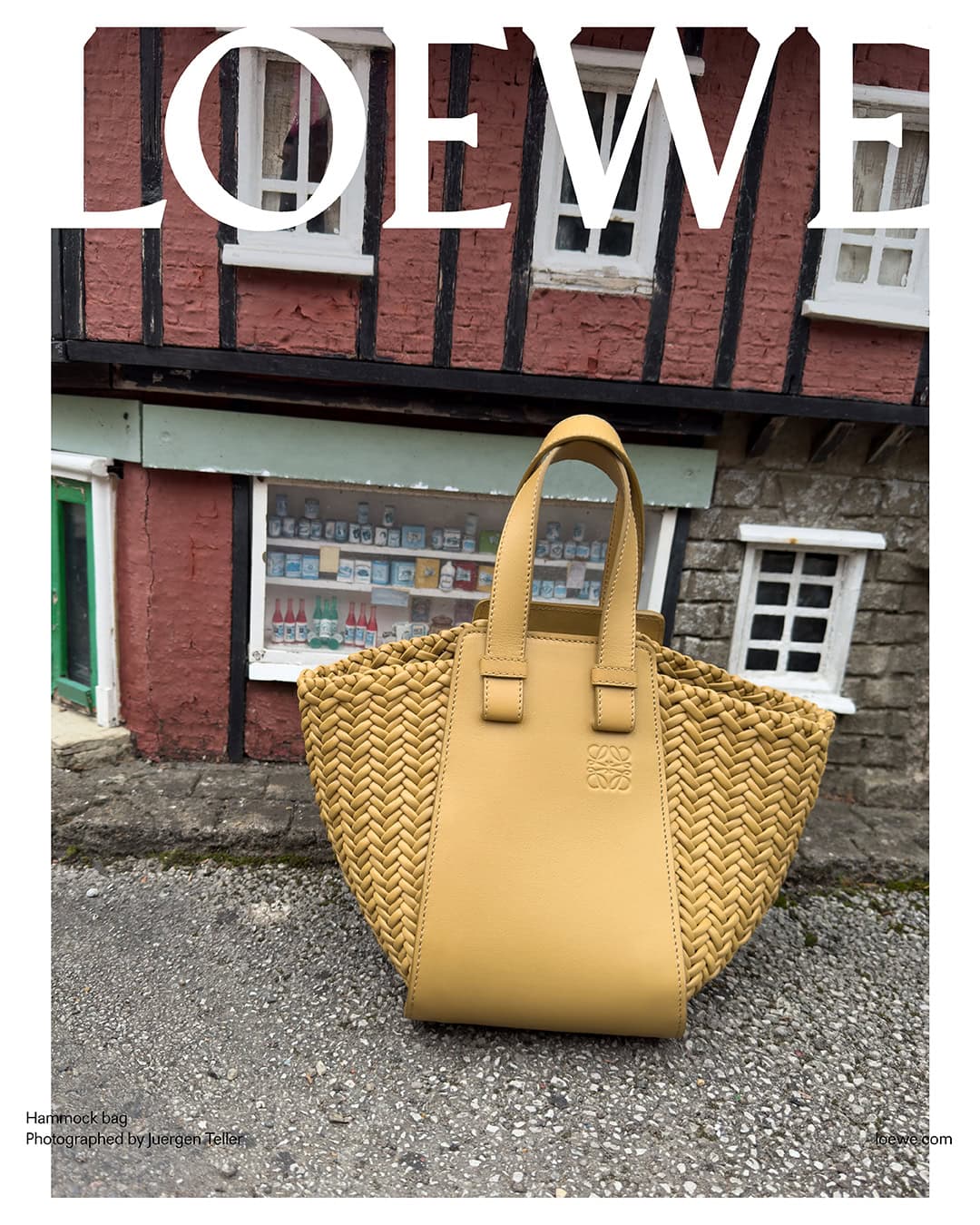 Tan leather and woven raffia Loewe Hammock bag showcased outdoors on a paved surface with a blurred building featuring small paned windows.