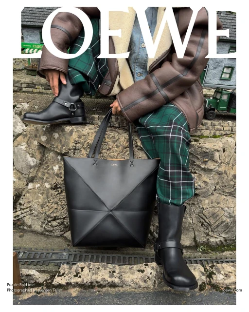 Fashion editorial featuring a person in green plaid pants and black boots, posing with a geometric black leather Loewe tote bag outdoors.