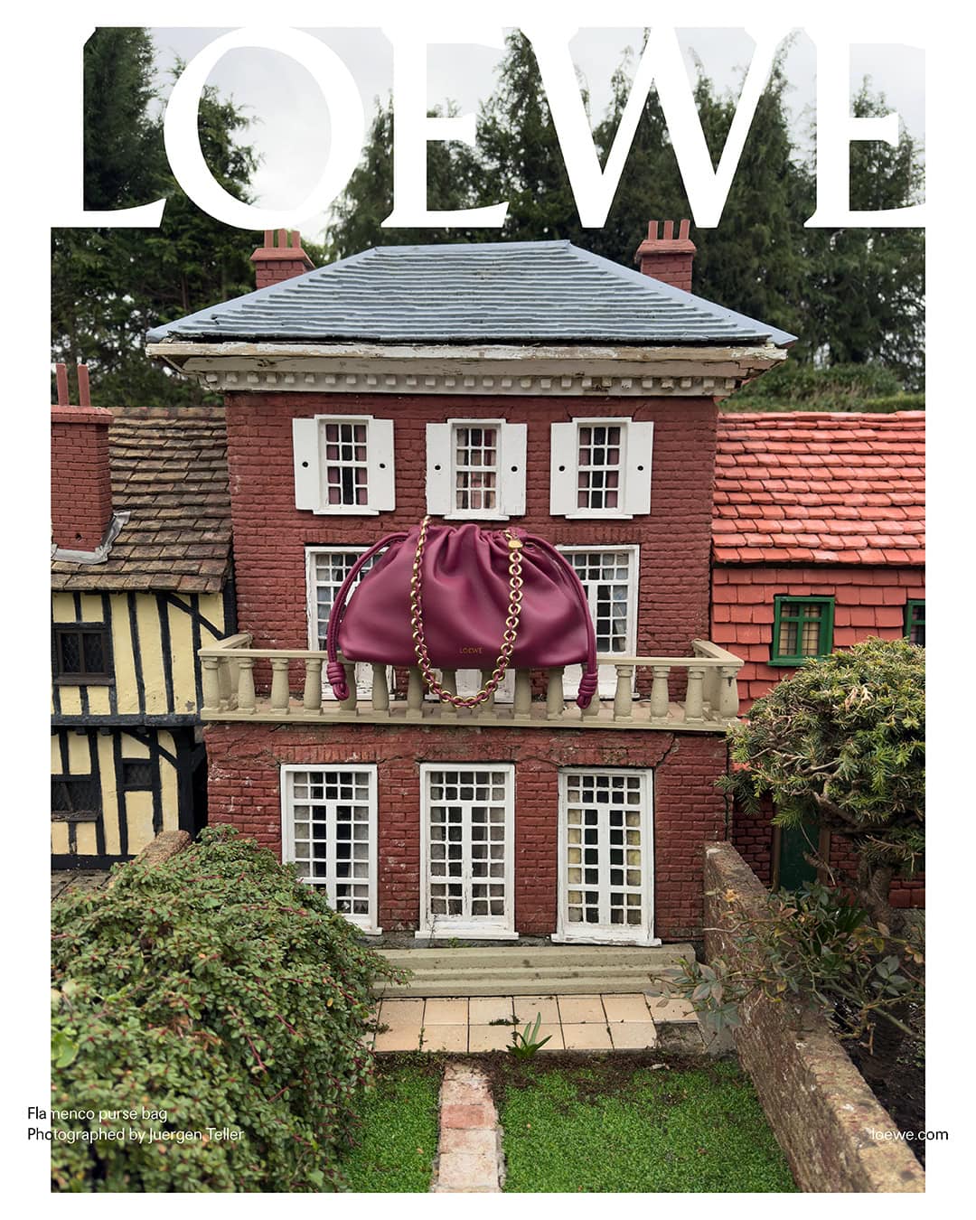 Burgundy leather Loewe bag with a gold chain on a brick house balcony with white windows, set in a miniature landscape with green bushes.