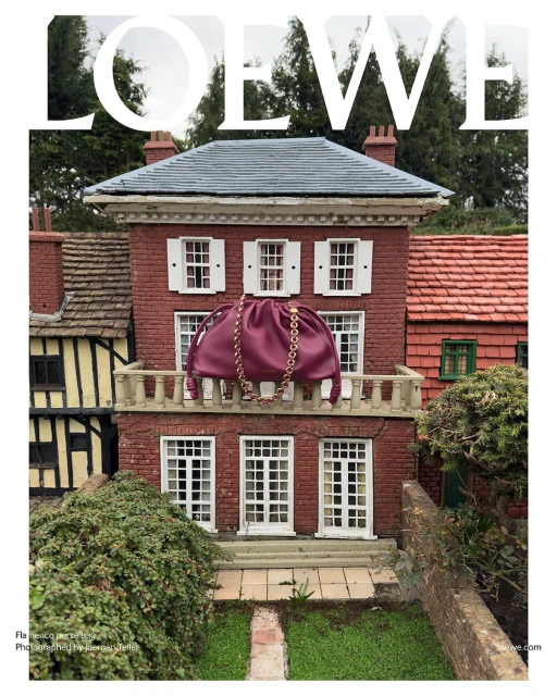 Burgundy leather Loewe bag with a gold chain on a brick house balcony with white windows, set in a miniature landscape with green bushes.